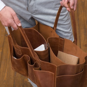 K&ouml;nnte beinhalten: Eine braune Ledertasche mit mehreren Taschen und F&auml;chern. Die Tasche hat zwei obere Henkel und ist offen, so dass ein wei&szlig;es Tablet und ein braunes Notizbuch sichtbar sind.