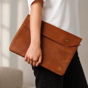 以下が含まれることがあります： 滑らかなマット仕上げの長方形の茶色のレザー製ノートパソコン用スリーブ。大きな封筒型のフラップと、中央に配置された真鍮色の金属製スナップボタンが特徴です。このスリーブは一枚の革から作られています。