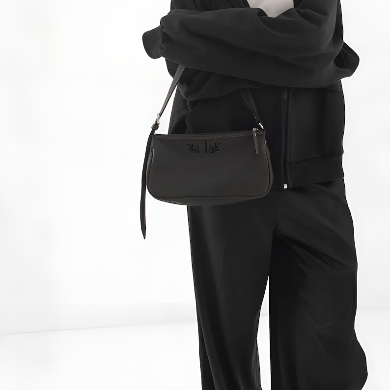 K&ouml;nnte beinhalten: Eine dunkelbraune Umh&auml;ngetasche mit langem Riemen. Die Tasche hat eine rechteckige Form mit abgerundeten Ecken und einem Rei&szlig;verschluss. Die Vorderseite der Tasche hat ein dekoratives Emblem. Die Person tr&auml;gt ein schwarzes Outfit.