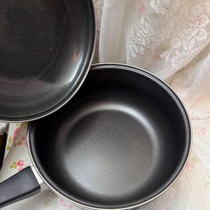 May include: A set of black non-stick cookware, including a saucepan with a black handle and a lid. The interior of the pan is a matte black, and the exterior is a shiny black. The cookware is set on a floral patterned fabric.