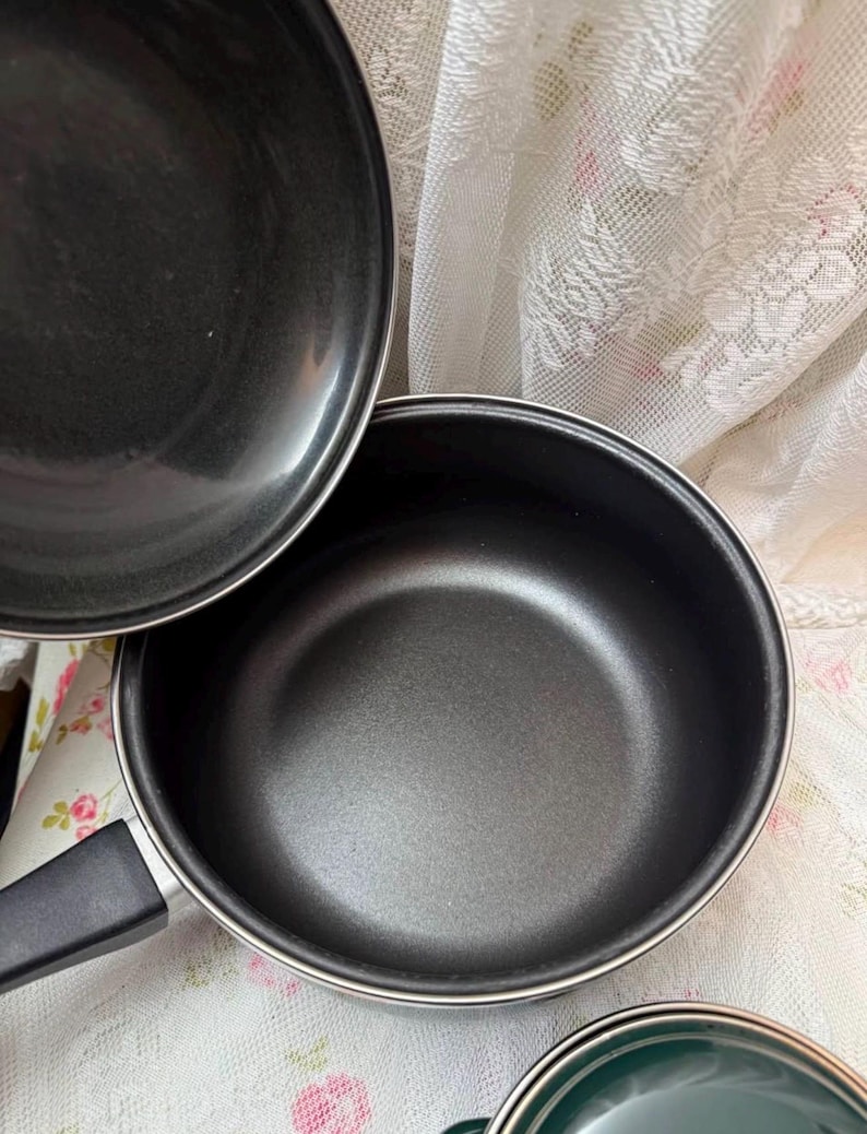 May include: A set of black non-stick cookware, including a saucepan with a black handle and a lid. The interior of the pan is a matte black, and the exterior is a shiny black. The cookware is set on a floral patterned fabric.