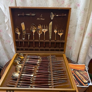 Può includere: Un set di posate dorate con manici in legno scuro, custodito in una valigetta di legno. Il set comprende vari cucchiai, forchette e coltelli, con il marchio Jean-Claude stampato in oro sulla fodera interna marrone del coperchio della valigetta.