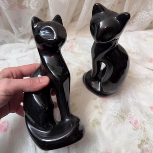 May include: Two glossy black cat ornaments. The cats are in a seated pose with their tails curled around their bodies. The ornaments have a sleek, minimalist design, with pointed ears and a smooth, reflective surface. The cats are displayed on a white lace surface.