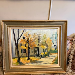 May include: An oil painting in a gold and green frame depicting a house surrounded by autumn trees with yellow and orange foliage. A path leads toward the building, and the artist's signature is visible in the bottom right corner of the canvas.