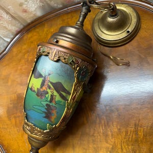 Może przedstawiać: Antyczna lampa wisząca w kolorze mosiądzu z namalowaną sceną krajobrazu. Lampa posiada cylindryczny klosz z kolorowym obrazem wiatraka i wody. Posiada dekoracyjne akcenty kwiatowe i liściaste oraz łańcuch do zawieszenia.