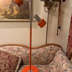 Puede incluir: Una lámpara de pie vintage naranja y cromada con dos focos ajustables. La lámpara tiene una base redonda y un tallo alto y delgado. Los focos son cilíndricos e inclinados. La lámpara está colocada frente a un sofá con estampado floral.