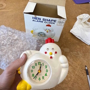 May include: A white hen-shaped alarm clock with a red comb and yellow feet. The clock face has black numbers and green hands. The box reads "HEN SHAPE ALARM CLOCK".