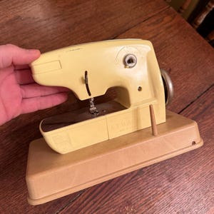 Può includere: Una macchina da cucire Straus vintage color crema. La macchina ha una parte superiore arrotondata con un quadrante e un ago. La base è di colore marrone chiaro. La macchina è tenuta da una mano.