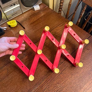 May include: A red plastic expandable coat rack with yellow knobs. The accordion-style rack is partially extended and held by a hand. The rack is on a wooden surface.