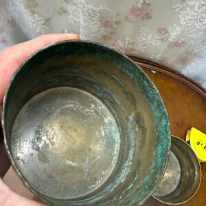 May include: A cylindrical metal container with a weathered, oxidized green patina on the exterior and a metallic silver-toned interior. A yellow measuring tape showing the text 2 x 6 is visible next to a smaller matching metal cup.