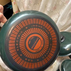May include: A set of green ceramic cookware with orange and black detailing. The base of the pan features the text "ELECTRIC", "INDUCTION", "GAS", "MEGAWARE", and "MADE IN SPAIN". The pan has a capacity of 2.8 liters.