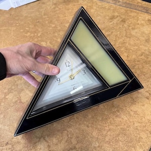 May include: A black and gold quartz clock in a triangular shape. The clock face is white with black numerals and gold hands. The clock has a gold trim and a light green accent.