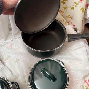 May include: A set of dark green saucepans with silver trim. The image shows a saucepan with a long handle and a lid being held open, plus two additional pots with lids. The lids and pots are dark green with silver trim.
