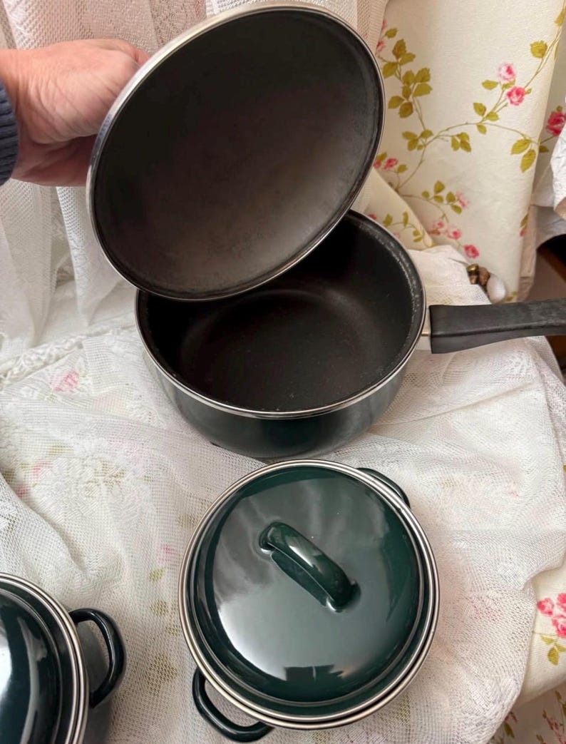 May include: A set of dark green saucepans with silver trim. The image shows a saucepan with a long handle and a lid being held open, plus two additional pots with lids. The lids and pots are dark green with silver trim.