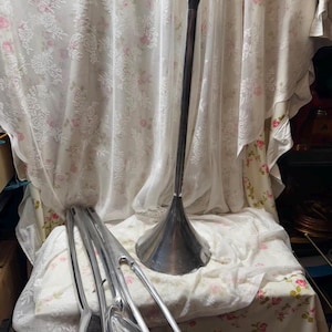 Puede incluir: Una lámpara de pie alta de metal plateado con una base acampanada y una parte superior de cuatro puntas. Varias piezas de metal plateado separadas están cerca. La lámpara está colocada sobre un fondo de tela con estampado floral.