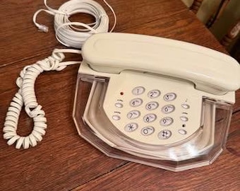 Może przedstawiać: Kremowy telefon przewodowy z przezroczystą plastikową podstawą. Telefon posiada standardową klawiaturę z cyframi i symbolami. Skręcony biały przewód łączy słuchawkę z podstawą. Inne akcesoria elektroniczne są widoczne w tle.
