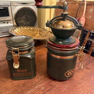 Peut inclure: Un ancien moulin à café avec une poignée en bois rouge et un corps vert foncé, à côté d'un pot assorti avec un fermoir doré. Le pot porte l'inscription "Harrods FOOD HALLS". Le moulin et le pot sont sur une surface en bois.