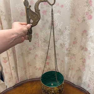 Pode incluir: Um vaso suspenso de latão com um suporte de parede em forma de dragão. O vaso tem um interior verde e uma faixa decorativa com figuras em relevo. O vaso é pendurado por uma corrente.