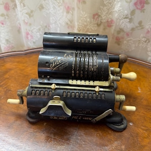 May include: A vintage black Thales mechanical calculator featuring a series of numbered rotating dials, metal levers, and white crank handles. The device displays the text Thales Patent and MEZ 46610 on its dark, metallic, multi-tiered body.