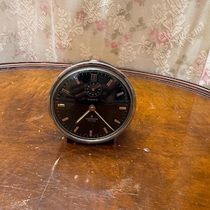May include: A round Junghans Trivox Silentic alarm clock with a chrome-finished metal frame and a black dial. The clock face features gold-toned hour markers, gold-toned hands, and a smaller sub-dial at the top. The text Junghans Trivox is printed.