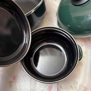 May include: A selection of dark green and black cooking pots and a lid. The pots have a shiny black interior and a silver rim. The lid has a black handle. The cookware is on a white floral patterned surface.