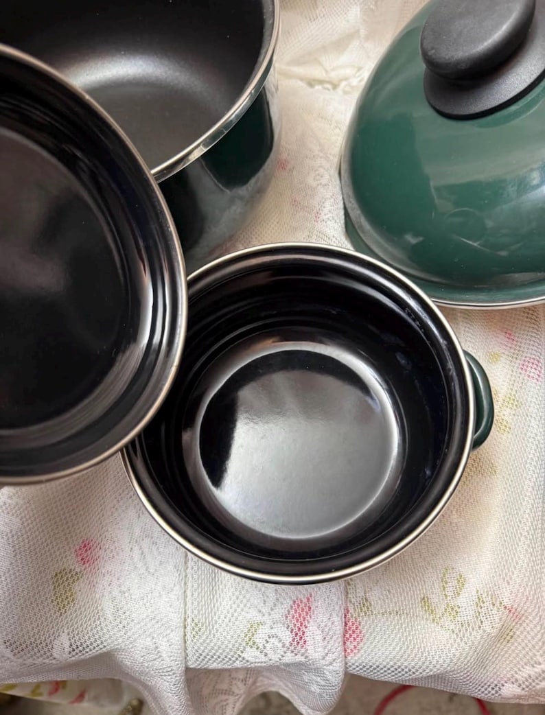 May include: A selection of dark green and black cooking pots and a lid. The pots have a shiny black interior and a silver rim. The lid has a black handle. The cookware is on a white floral patterned surface.
