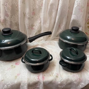 May include: A collection of four dark green saucepans and pots with black handles and lids. The set includes two saucepans with handles, and two smaller pots with loop handles. The cookware is displayed on a floral patterned surface.