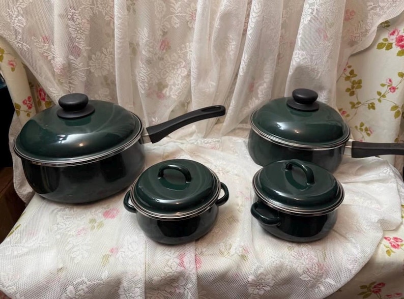 May include: A collection of four dark green saucepans and pots with black handles and lids. The set includes two saucepans with handles, and two smaller pots with loop handles. The cookware is displayed on a floral patterned surface.