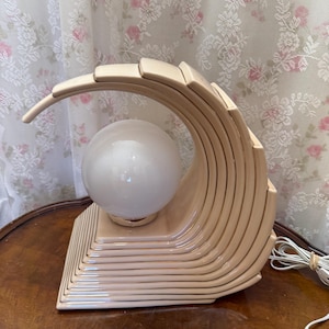 May include: A beige ceramic table lamp featuring a curved, stepped wave-like structure that arches over a central, spherical white glass globe light bulb. The base is rectangular with tiered, horizontal ridges that follow the curve of the lamp.