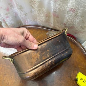 May include: An oval copper basin with a dark, weathered patina and two brass handles on the sides. The basin features a horizontal ridge detail along the upper body. A yellow measuring tape is visible nearby, showing a 6-inch measurement mark.