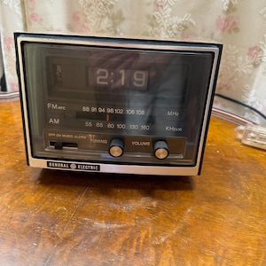 Può includere: Una radiosveglia digitale rettangolare General Electric con frontalino nero, bordo argentato e display digitale che mostra 2:19. Il dispositivo è dotato di scale di frequenza radio AM e FM, una manopola di sintonia, una manopola del volume e un interruttore a scorrimento.