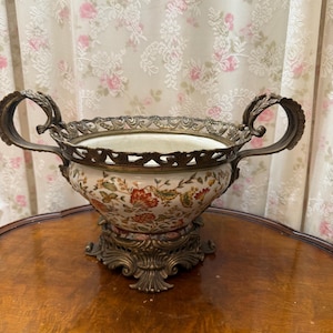 May include: A ceramic bowl featuring a colorful floral pattern with red, orange, and green tones. The bowl is set within an ornate, dark metal frame with two curved handles and a decorative base, featuring intricate scrollwork and filigree details.