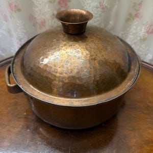 Puede incluir: Una olla redonda de cobre martillado con una tapa abovedada a juego. La tapa presenta un asa central circular y acampanada. La olla tiene una pátina oscura y desgastada, e incluye un asa lateral visible a la izquierda. La superficie es irregular.