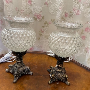 Op de afbeelding: Twee vintage tafellampen met heldere, diamantpatroon glazen kappen en sierlijke, bronskleurige voetstukken. De lampen hebben een gegolfde rand aan de bovenkant van de kap en staan op een houten tafel. Witte snoeren zijn zichtbaar.