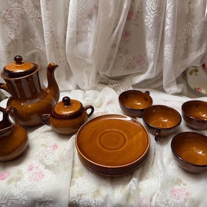 Pode incluir: Um conjunto de chá vintage em cerâmica marrom, incluindo uma chaleira, um creme, um açucareiro, pires e xícaras. O conjunto tem um acabamento brilhante e é disposto sobre uma toalha de mesa de renda branca. Design clássico.