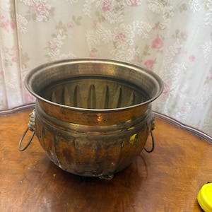 May include: A vintage brass planter with a round body and a wide rim. The planter features decorative lion head handles and a ribbed design on the lower portion. The interior shows signs of age and use, adding to its antique charm.