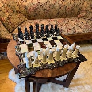 Puede incluir: Un juego de ajedrez decorativo con piezas blancas y negras sobre un tablero a cuadros. El tablero está enmarcado en plata y se encuentra sobre una mesa redonda de madera. El juego de ajedrez se exhibe sobre una mesa pequeña de color marrón oscuro.