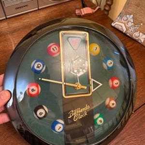 Könnte beinhalten: Runde Uhr mit schwarzem Rahmen und grünem Zifferblatt, auf dem kleine Billardkugeln als Stundenmarkierungen dienen. Die Uhr hat einen goldfarbenen Rahmen in der Mitte und trägt die Aufschrift "Billiards Clock". Die Zeiger der Uhr sind goldfarben.