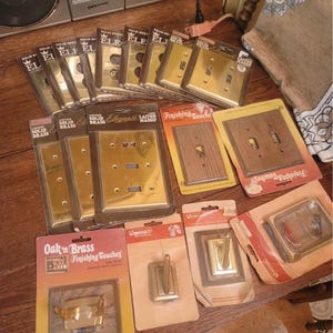 May include: A selection of vintage brass electrical switch plates, outlet covers, and hooks, some still in their original packaging. The packaging features text like "Solid Brass" and "Finishing Touches". The items are arranged on a wooden surface.