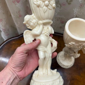 Puede incluir: Un cáliz decorativo de color crema que presenta una figura de querubín esculpida sosteniendo una copa estriada adornada con motivos florales y de frutas en relieve. La base del cáliz es circular e incluye un patrón decorativo repetitivo.