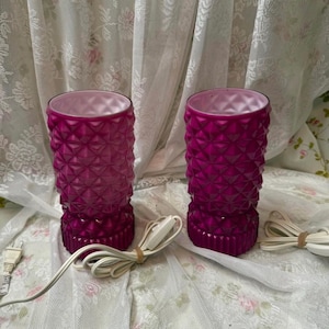 May include: Two vintage magenta glass lamps with a textured, geometric pattern. Each lamp has a cylindrical shape with a flared base and a matching magenta interior. White electrical cords are visible.