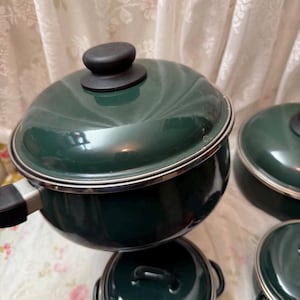May include: A collection of dark green enamel cooking pots and pans with black handles and lids. The cookware features a silver trim and is displayed on a floral patterned surface. The set includes a saucepan, a pot, and two smaller pans.