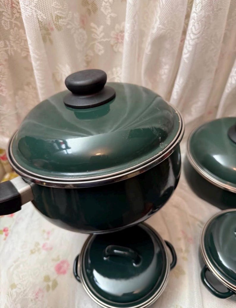 May include: A collection of dark green enamel cooking pots and pans with black handles and lids. The cookware features a silver trim and is displayed on a floral patterned surface. The set includes a saucepan, a pot, and two smaller pans.