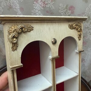 May include: A small, decorative wooden shelf unit with a cream-coloured finish and gold accents. The unit features arched openings, shelves, and ornate corner details. The interior is lined with red fabric.