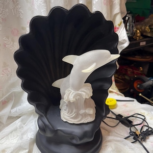Puede incluir: Lámpara negra en forma de concha con una escultura de delfín blanco. El delfín salta sobre una ola. La lámpara tiene un borde festoneado y una base redonda. La lámpara está hecha de un material mate.