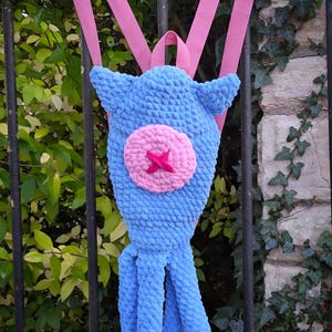 Puede incluir: Una mochila azul tejida a crochet con forma de pulpo, con correas rosas. Tiene un círculo rosa con una estrella rosa. La mochila está colgada de una valla metálica negra, con follaje en el fondo.