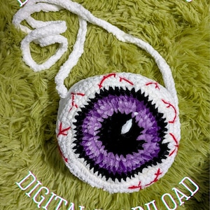May include: A white, round, crocheted eye-shaped purse with a purple iris and black pupil. Red veins are stitched around the eye. The purse has a white strap. The words "DIGITAL DOWNLOAD" are in the background.