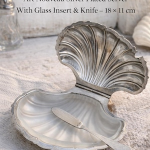 Peut inclure: Un beurrier antique en forme de coquillage argenté avec un insert en verre et un couteau. Le plat, de 18 x 11 cm, présente un design Art Nouveau. Le couvercle est ouvert, révélant l'intérieur et le petit couteau à beurre.