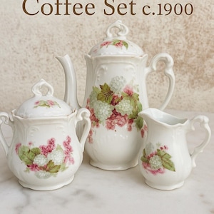 Op de afbeelding: Een vintage witte porseleinen koffieset, bestaande uit een theepot, suikerpot en melkkan. Elk stuk is versierd met een bloemmotief in roze, groen en wit. De set dateert van rond 1900.