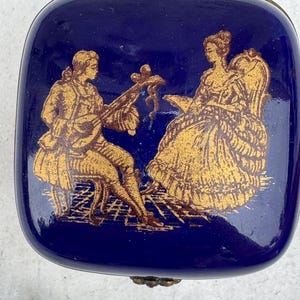 Puede incluir: Caja de baratijas rectangular azul marino con ribete met&aacute;lico dorado. La tapa presenta una ilustraci&oacute;n dorada de un hombre tocando un instrumento de cuerda y una mujer sentada en una silla.